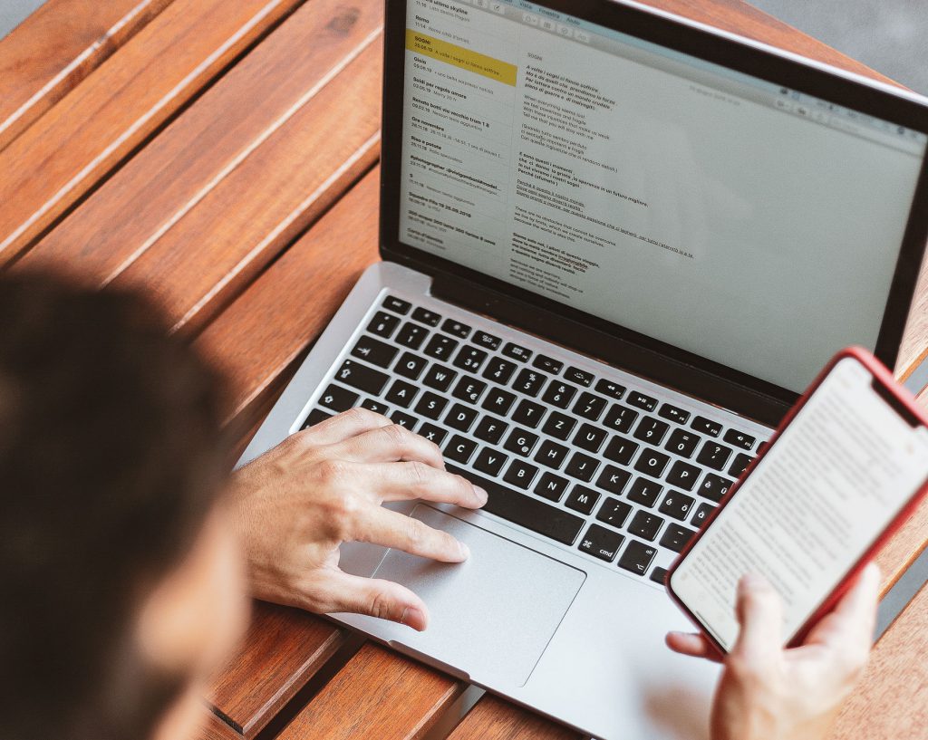 Over head shot of someone using laptop on top of a wooden table whilst looking at phone screen