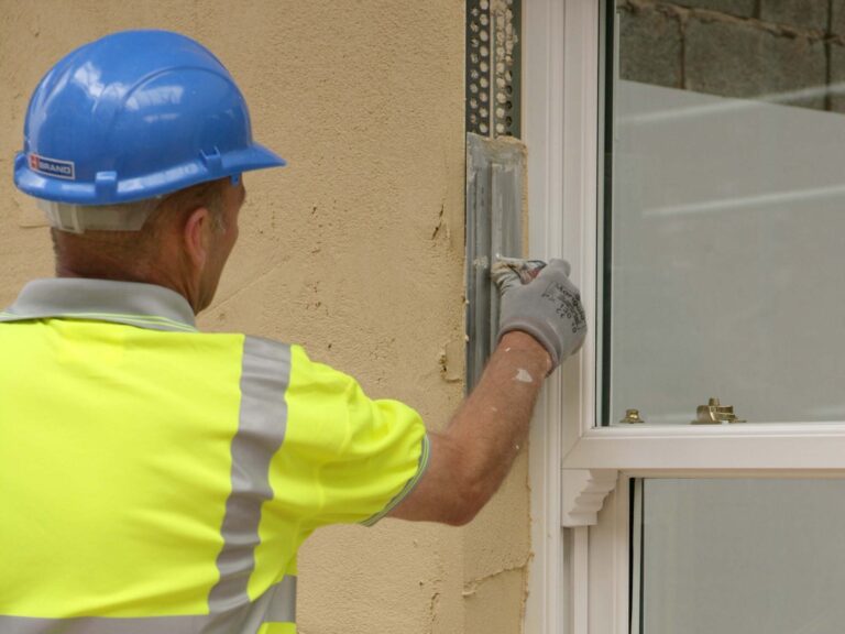 Construction worker with high vis, hard hat and gloves applying render on top of PVCu render beading at window reveal