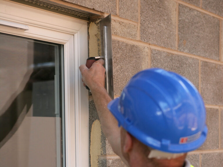Construction worker with blue hard hat applying render dabs to window reveal on block work